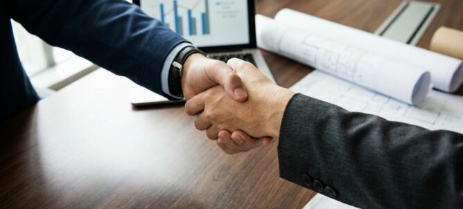 Zakelijke handdruk boven houten conferentietafel met laptop, grafieken en technische documenten op achtergrond